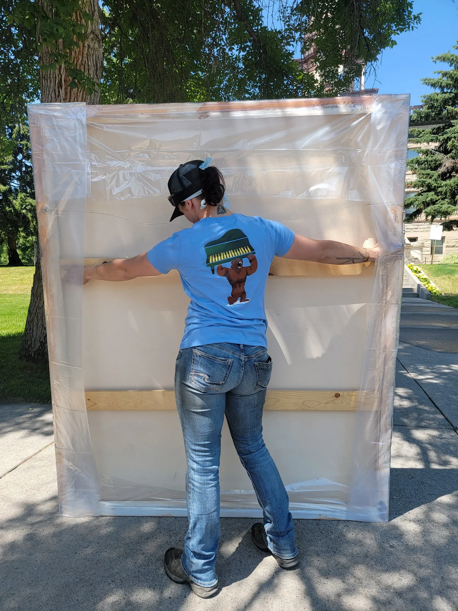 A Moving On Up crew member in a branded bear-and-piano logo shirt shrink-wrapping a mattress outdoors