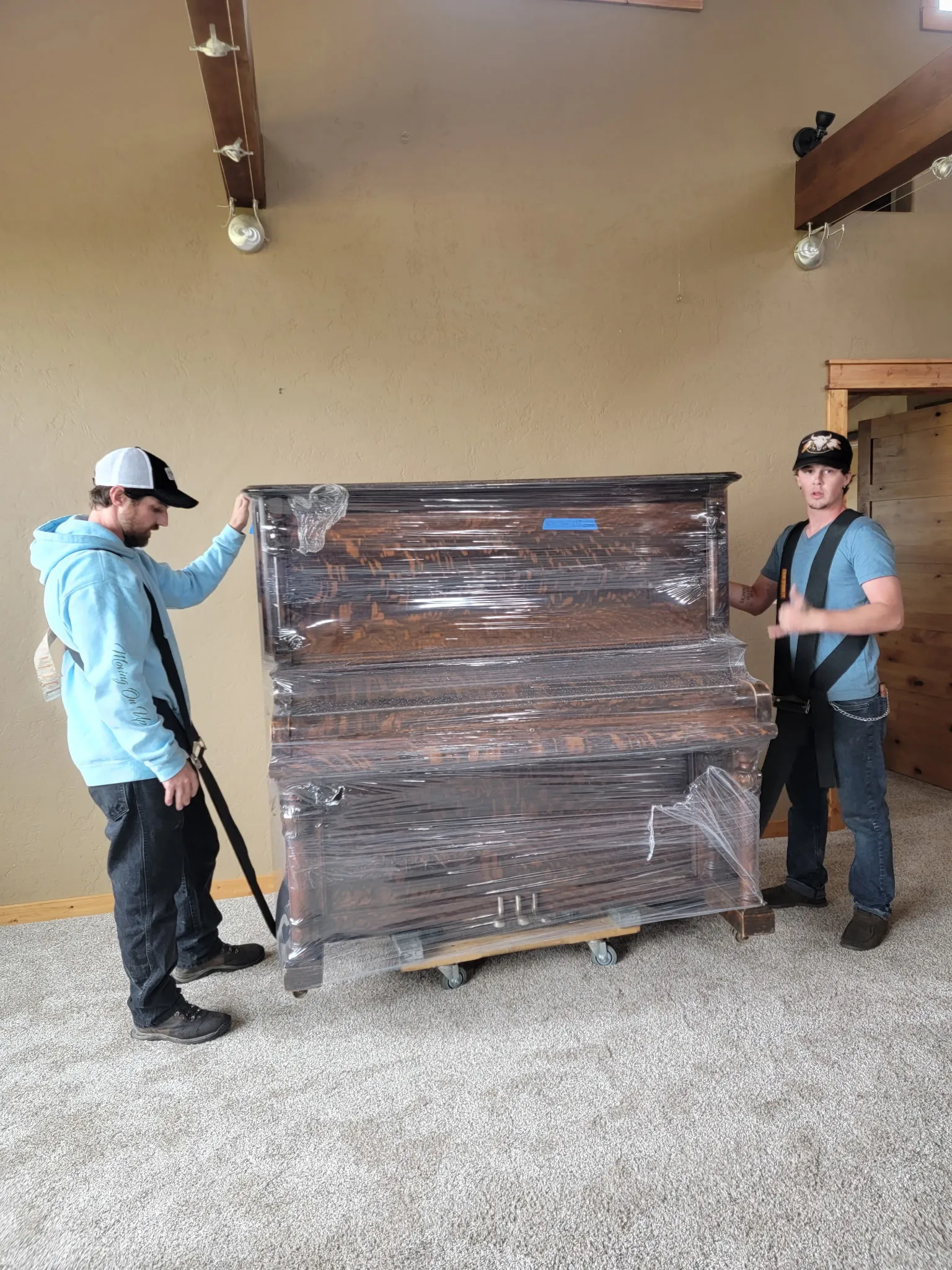 Two Moving On Up crew members rolling a shrink-wrapped upright piano on a dolly inside a home