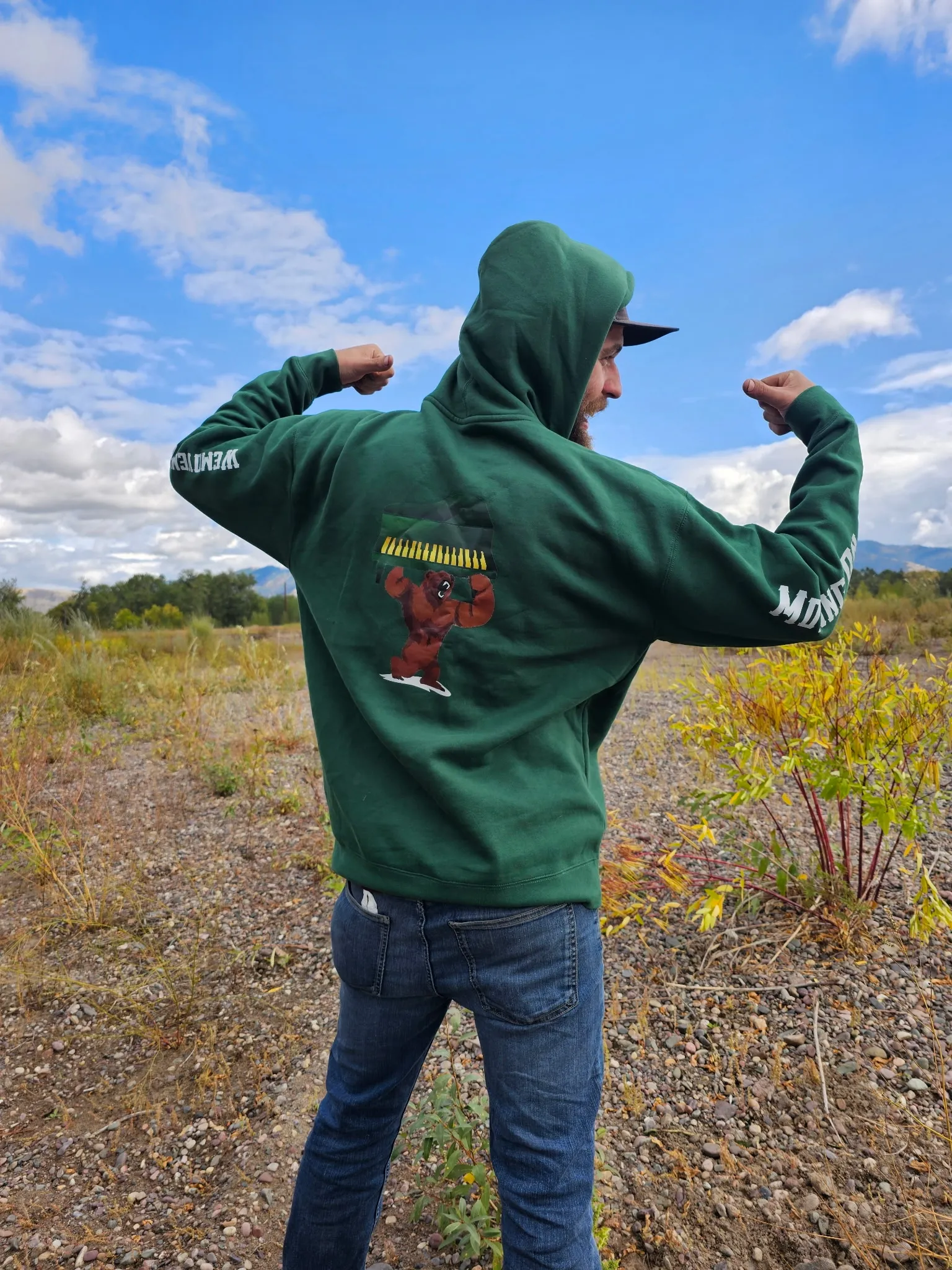 A Moving On Up team member in a green hoodie showing the grizzly-and-piano logo on the back, flexing in a western Montana field